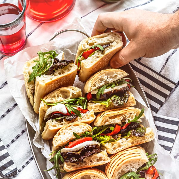 A hand reaching for a SUPPA'S PIZZA & SUBS sub sandwich with red peppers, mozzarella, and arugula.