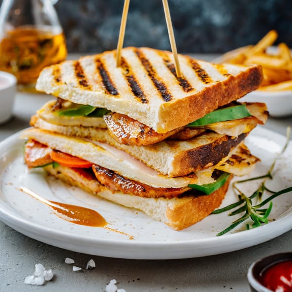A stacked grilled chicken sandwich with vegetables and cheese, served with fries and sauce.