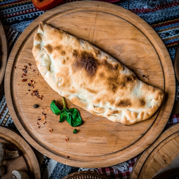 A golden-brown calzone with basil and spices on a round wooden board.
